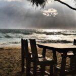 Wooden table and chairs on a sandy beach facing the sea under dramatic sunlight – nbcreations