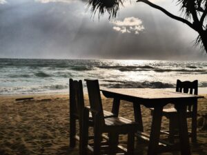 Wooden table and chairs on a sandy beach facing the sea under dramatic sunlight – nbcreations