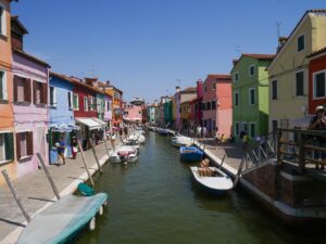 Canal in Murano lined with colorful houses and moored boats, and poeple walking – Venetian island scene - nbcreations