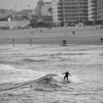 Black and white side view of a surfer riding a wave toward the beach – minimalist surf scene - nbcreations