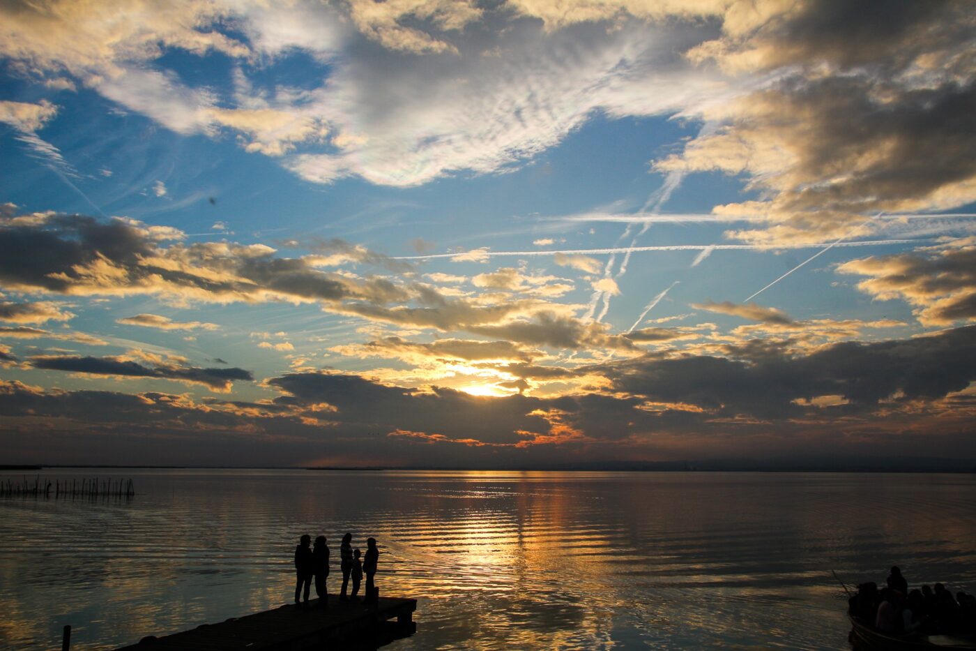 Silhouettes of people standing on a pier at sunset, with dramatic clouds and sky reflections on a calm lake – nbcreations