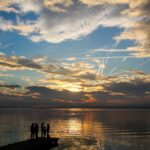 Silhouettes of people standing on a pier at sunset, with dramatic clouds and sky reflections on a calm lake – nbcreations
