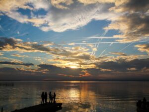 Silhouettes of people standing on a pier at sunset, with dramatic clouds and sky reflections on a calm lake – nbcreations