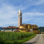 Church with a tall bell tower in a rural Italian village, surrounded by cornfields under a blue sky. - nbcreations